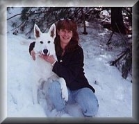 An American White Shepherd is sitting next to a lady in a snowy yard. the Shepherd has its paw on the leg of the lady next to it.