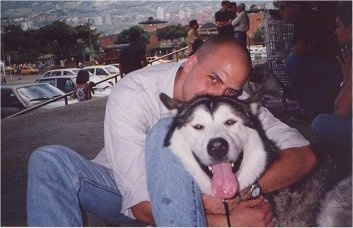 A black and white Alaskan Malamute is sitting with a crowd being hugged by a person with its tongue out and mouth open