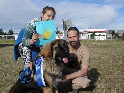 Kabrone the Caucasian Ovtcharka ihas a blue ribbon on it. It is sitting in front of his owner and a girl. The Girl behind Kabrone is holding a piece of cardboard with a drawing on it