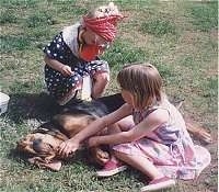 A girl in a blue polka dot dress and a red bandana is kneeling behind a sleeping Bloodhound. In front of it is a girl in a pink dress sitting in dirt.