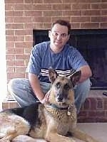 A black and tan German Shepherd is laying in front of a person sitting in front of a brick fireplace