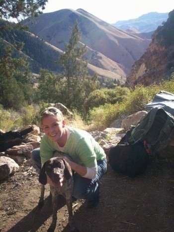 A white with brown German Shorthaired Pointer is standing in a dirt path with a lady behind it. There is a backpack to the left of them