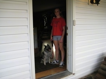 A blonde-haired girl is standing in a doorway next to a black, grey and white Norwegian Elkhound dog that is sitting.
