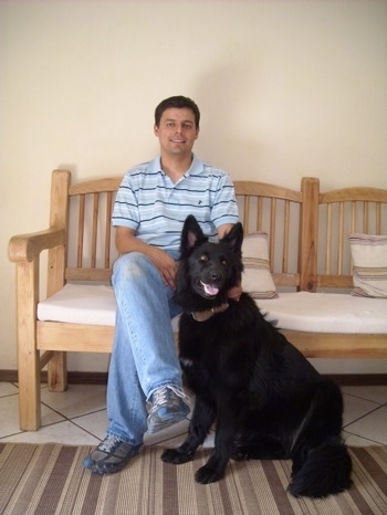 A man is sitting on a wooden bench inside of a room and there is a black German Shepherd sitting in front of it