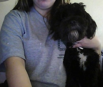 A black with white Malti-Pug dog is sitting next to a lady who has her arm around the side of a dogs body.