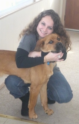 A kneeling lady in a gray sweater is hugging a big red dog that is standing over her knee and they both are looking forward.