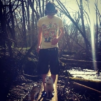 A white American Eskimo puppy is sitting on a log with a person behind him standing on the log with muddy water around them