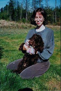 A brown Spanish Water Dog puppy is being held against the body of a lady taking a knee in a yard. The puppy has a rope toy in its mouth.