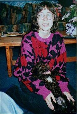 A lady in a pink with black and red Sweater is holding a brown with white Spanish Water Dog puppy against her lap. the puppys mouth is open and it is looking up.