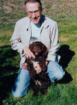 A man in a tan jacket is taking a knee in a yard and he is holding a brown with white Spanish Water Dog puppy up against his lap.