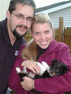 A lady in maroon and a man in black are standing next to each for a photo while the lady is holding a tiny black with white and tan Shetland Sheepdog in her arms. Everyone is looking forward.