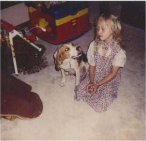 Bugle the Briquet licking his nose and sitting next to a kneeling little girl with baby toys next to them