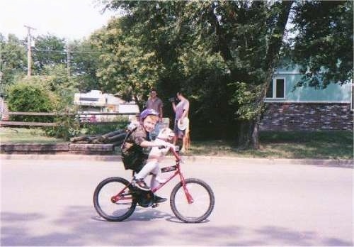 Sally the Poodle mix is riding down the street on a bike with a boy named Tyler. The dog is in front of the boy sitting on the seat with its paws on the handle bars.