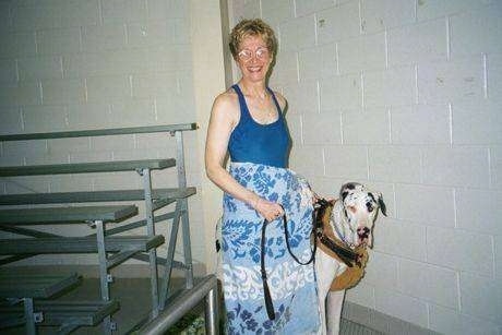 Baily the Great Dane and Dale are standing in front of a brick wall and there is a metal bench behind them at an indoor pool.