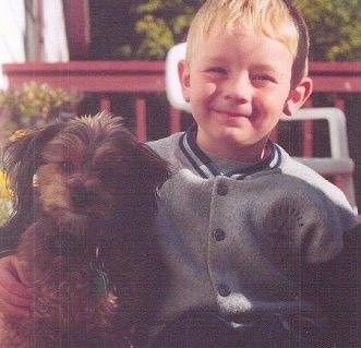 A boy in a letterman jacket is sitting on a chair next to a brown with black Pomapoo puppy. They are looking forward sitting on a porch.