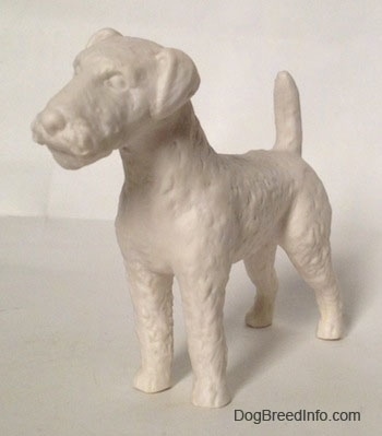 The front left side of a white bisque porcelain Airedale Terrier dog figurine that is unglazed. The dog has straight legs and a straight muzzle.