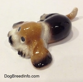 The front left side of a black and tan with white ceramic Basset Hound figurine that is laying down. The figurine is glossy.