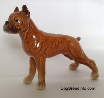 The left side of a porcelain brown Boxer dog figurine in a show stance. The figurine has a black circles for eyes.