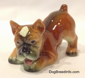 The front left side of a brown, black and white Boxer puppy figurine in a play bow pose. The figurines mouth is open.