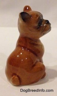 The back right side of a brown with white Boxer puppy figurine that is in a sitting pose. The figurine has a short tail.
