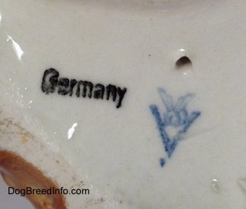 The underside of a porcelain brown with white and black Boxer dog figurine. There is the Goerbel W. Germany logo, above it is a hole and to the left of it is a stamp of the word - Germany.