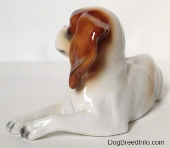 The front left side of a white with brown and black Cavalier King Charles Spaniel figurine that is laying down. The tips of the figurines paws are black.