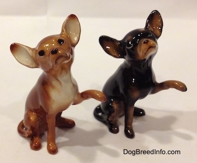 Two different Chihuahua figurines that have one paw in the air. The figurines are glossy.