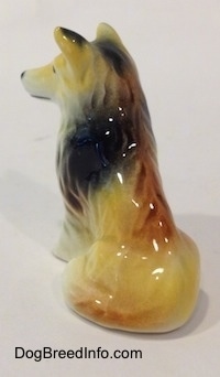 The back left side of a tan, black and white rough coated Collie dog that is in a sitting pose. The back of the figurine is glossy.