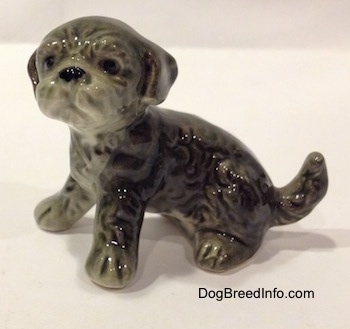The left side of a black and gray Dandie Dinmont Terrier puppy in a sitting position figurine. The figurine has fine hair details and it has black circles for eyes. Its nose is black and its ears hang down to the sides of its head. Its snout is pushed back.