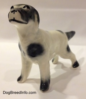 The front right side of a ceramic figurine of a white and black English Setter in a standing pose. The figurine has lines for its mouth and black circles for its eyes.