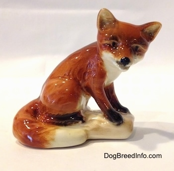 The right side of a porcelain red fox figurine sitting on a log. The figurine has black circles for eyes and a black nose.
