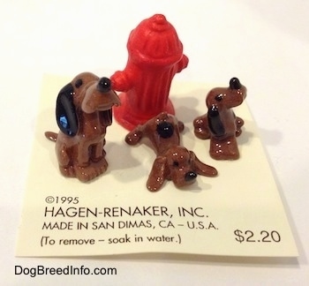 A set of Hound Dawg figurines that are around a red fire hydrant. One of the figurines is sitting, another is laying down and another is looking up and to the right.