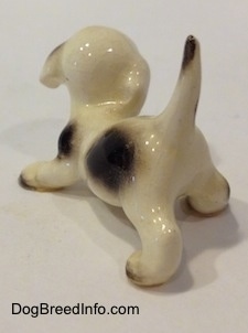 The back left side of a white with brown Hound puppy crouching figurine. The figurine has a long tail sticking up in the air and it has a brown spot at the tip.