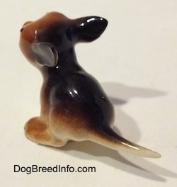 The back left side of a figurine of a miniature running puppy. The figurine has a long skinny tail.