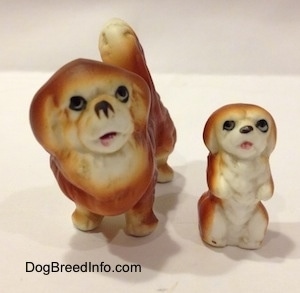 A brown with white Pekingese puppy figurine that is in a begging pose and to the left of it is a brown with white Pekingese figurine. The figurines mouthes are open.