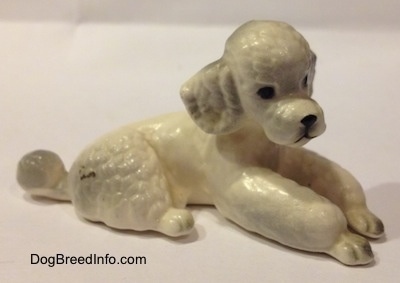 The front right side of a ceramic figurine of a white Poodle that is in a lying position. The figurine has poofy legs.