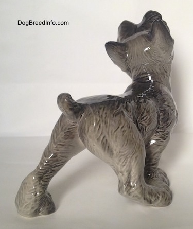 The back right side of a grey with white Schnauzer puppy figurine that is walking. The figurines tail is arched up.