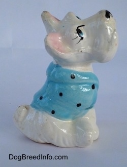 The right side of a white ceramic Scottish Terrier sitting and it has a blue with black shirt on figurine. The figurine has small perky ears.