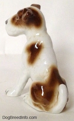 The back left side of a white with brown figurine of a Wire Fox Terrier in a sitting position. The figurine has a short tail that is curled onto its back.