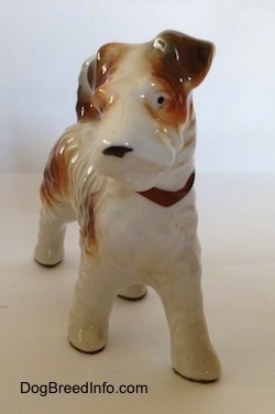 A white with tan figurine of a Wire Fox Terrier standing. The figurine has small black dots for eyes.