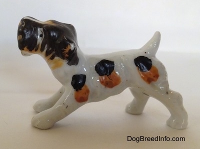 The left side of a white with black and brown bone china Wire Fox Terrier in a play bow pose figurine. The figurine has black and brown spots along its body.