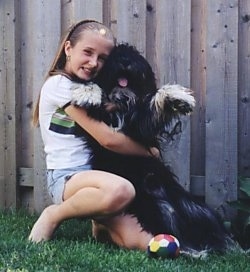 A blonde haired girl is hugging a black with white Polish Lowland Sheepdog.