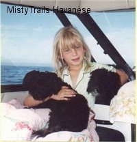A blonde-haired girl is sitting in a boat and she is holding onto two black dogs. They are all looking forward.