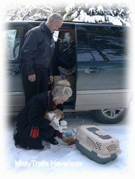 A blonde-haired lady is taking a puppy out of a crate. A man in a black leather coat is standing behind her.