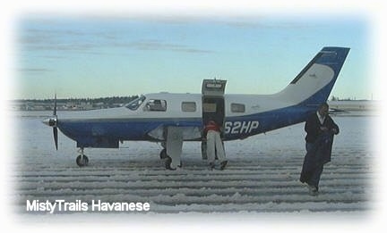 The left side of a private plane on a runway that has a small amount of snow all over it. There is a person walking away from the plane.