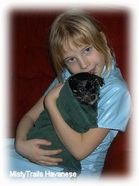 A blonde-haired girl is holding a wet black puppy wrapped in a green towel, close to her.