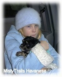 A girl in a fuzzy hat and a blue jacket is sitting in the back of a vehicle and she is holding a black with gray Havanese puppy as she crys.