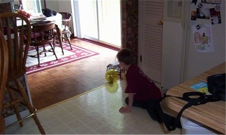 A hamster is rolling around in a yellow exercise ball inside of a house and next to it is a boy laying on a tiled floor.