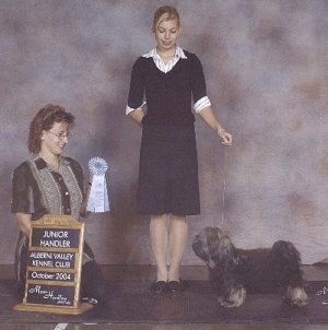 A blonde haired girl is standing up for a photo, in front of her is a lady holding a ribbon and to the right of her is a dog that is posing.