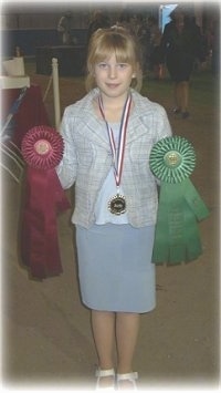 A blonde-haired girl is holding a red ribbon in her left hand, a green ribbon in her right hand and she has a medal around her neck.